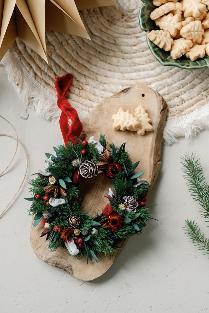 Decorative mini dried flower wreath bauble on a wooden board with cookies and a red ribbon.