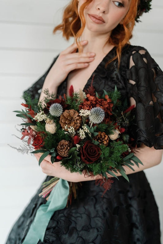 A traditional festive winter bridal bouquet featuring preserved red roses, thistles and lush greenery, dramatic and full of character, finished with green silk ribbon by Luna and Wild.  