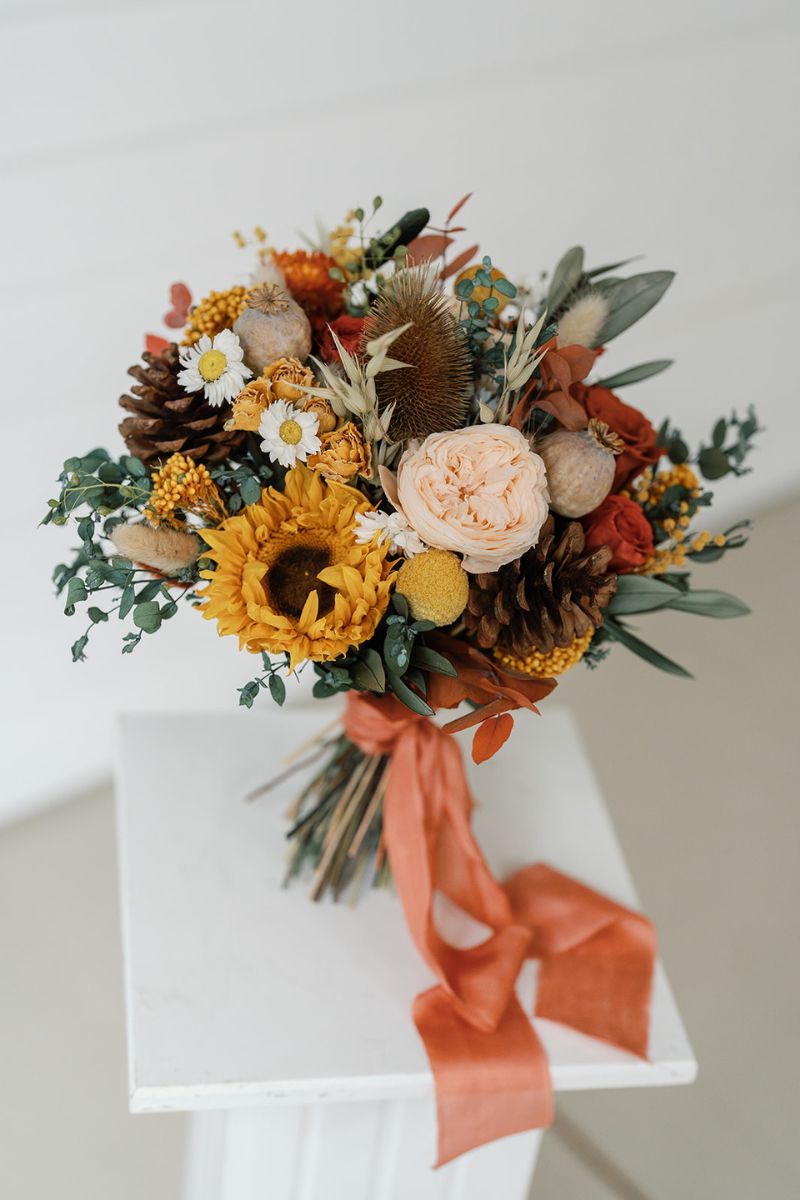 autumnal themed dried flower bridal bouquet in shades of mustard yellow, rust, crowns and lush greenery, hand tied with a rust silk ribbon.