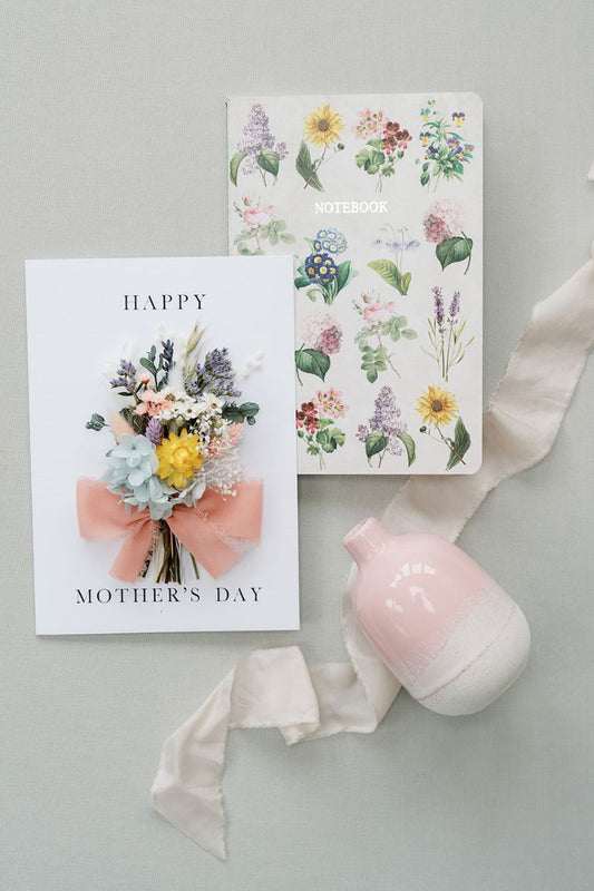Mothers day gift bundle containing a dried flower happy mother card card tied with a ribbon bow, a pink and ivory dip dye mini vase and wildflower design notebook.