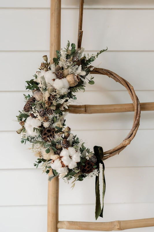 Decorative wreath with cotton, pinecones, and greenery on a wooden hoop against a white wall.